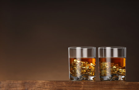Whiskey with ice cubes in glasses on wooden table against brown background, space for textの写真素材