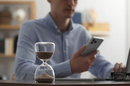 Hourglass with flowing sand on desk. Man using smartphone indoors, selective focusの写真素材