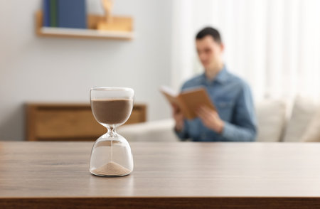 Hourglass with flowing sand on desk. Man reading book in room, selective focus