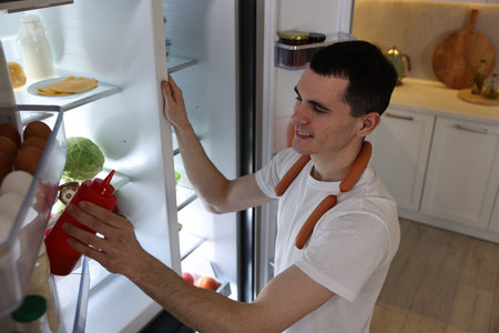 Happy man with sausages taking ketchup out of refrigerator in kitchenの写真素材