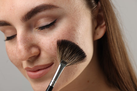 Beautiful woman with freckles applying makeup with brush on gray background, closeup