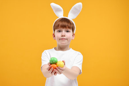Easter celebration. Cute little boy with bunny ears and painted eggs on orange backgroundの写真素材