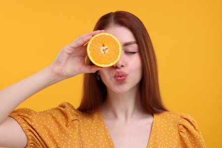 Beautiful woman covering eye with half of orange and sending air kiss on yellow backgroundの写真素材