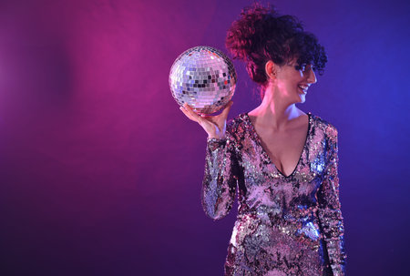 Beautiful young woman with disco ball posing on color background in neon lights. Space for text