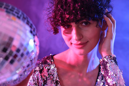 Beautiful young woman with disco ball posing on color background in neon lightsの写真素材