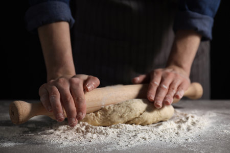Making bread. Woman rolling dough at table on dark background, closeup