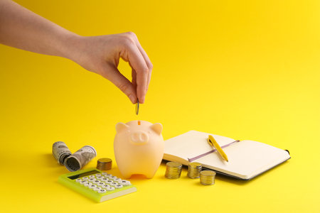 Financial savings. Woman putting coin into piggy bank on yellow background, closeupの写真素材