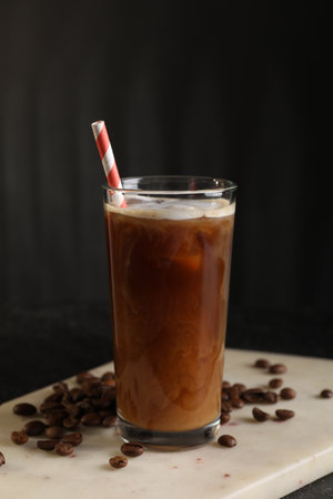 Refreshing iced coffee with milk in glass and beans on table against dark gray background