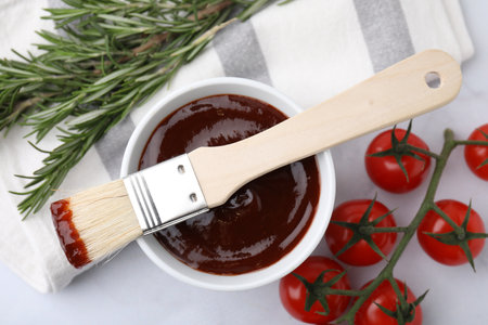 Marinade in bowl, basting brush, tomatoes and rosemary on white table, flat layの写真素材