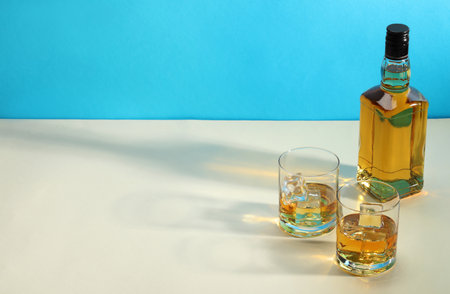 Whiskey with ice cubes in glasses and bottle on white table against light blue background, space for textの写真素材