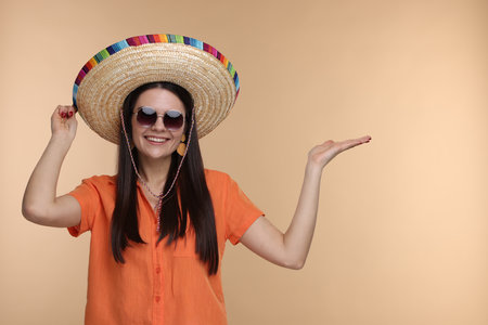 Young woman in Mexican sombrero hat and sunglasses showing something on beige background. Space for textの写真素材