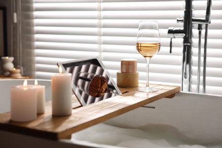 Wooden tray with tablet, wine and candles on bathtub in bathroom