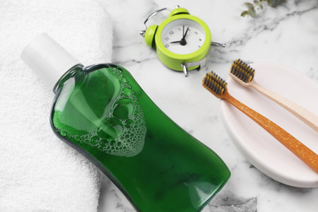 Fresh mouthwash in bottle, toothbrushes and alarm clock on white marble table, above viewの写真素材