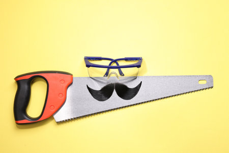 Man's face made of artificial mustache, safety glasses and hand saw on yellow background, top viewの写真素材