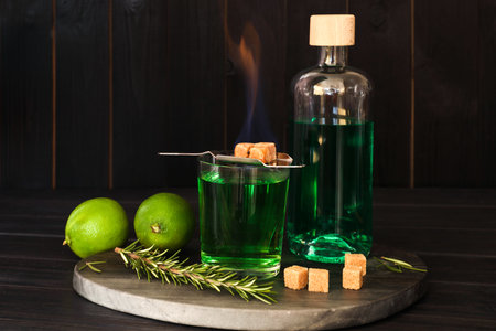 Absinthe in glass, flaming brown sugar, rosemary and lime on wooden table. Alcoholic drink