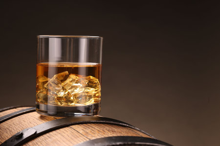 Whiskey with ice cubes in glass on wooden barrel against dark background, closeup. Space for textの写真素材