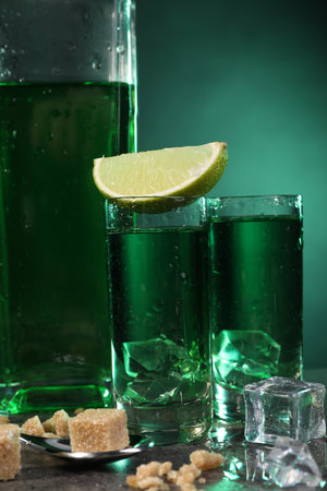Absinthe in shot glasses with ice cubes, lime, brown sugar and spoon on gray table, closeup....