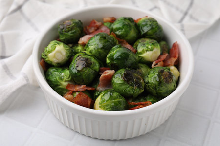 Delicious roasted Brussels sprouts and bacon in bowl on white tiled table, closeupの写真素材