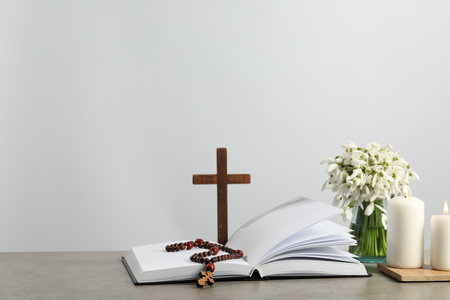 Church candles, Bible, wooden cross, rosary beads and flowers on gray table. Space for text