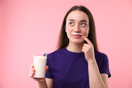 Cute woman with milk mustache holding glass of tasty dairy drink on pink backgroundの写真素材