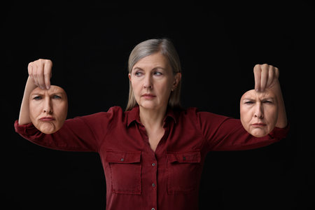 Mature woman holding masks with his face showing different emotions on black background. Balanced personalityの写真素材