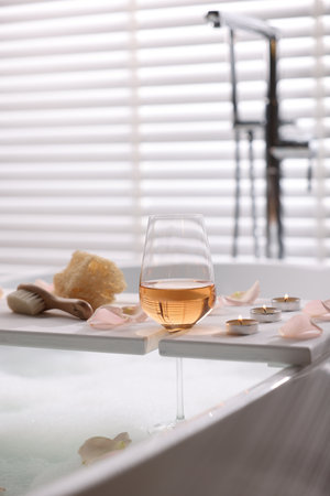 Wooden tray with wine, toiletries and flower petals on bathtub in bathroomの写真素材
