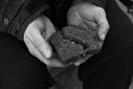 Poor homeless man holding piece of bread outdoors, closeup. Black and white effect