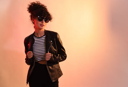 Beautiful young woman in black leather jacket and sunglasses posing on color background in neon...