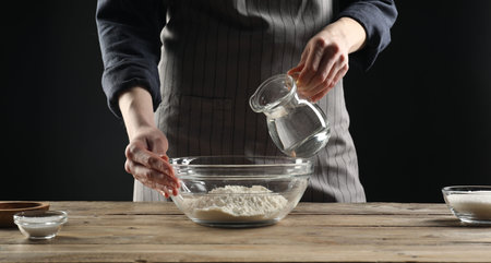Making bread. Woman pouring water into bowl with flour at wooden table, closeupの写真素材