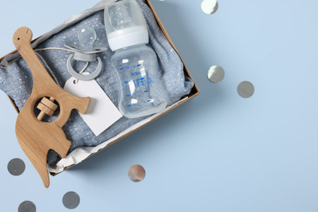 Different baby accessories with blank card in box and confetti on light blue background, top view....