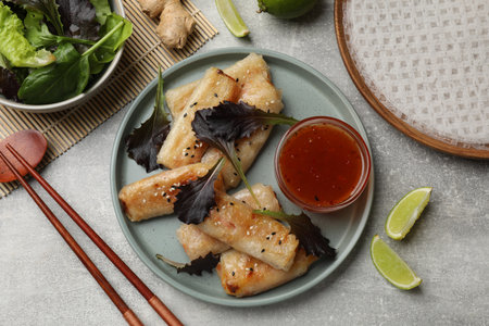 Tasty fried spring rolls, lettuce, lime and sauce on gray table, flat lay
