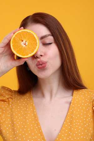 Beautiful woman covering eye with half of orange and sending air kiss on yellow backgroundの写真素材
