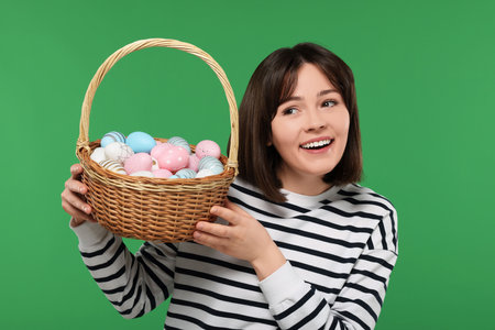 Easter celebration. Happy woman with wicker basket full of painted eggs on green backgroundの写真素材