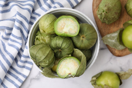 Fresh green tomatillos with husk in bowl on light marble table, flat lay