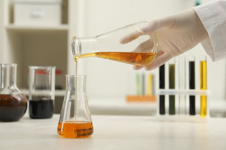 Laboratory worker pouring orange crude oil into flask at light marble table, closeup