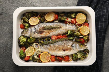 Delicious fish with vegetables and lemon in baking dish on gray textured table, top view