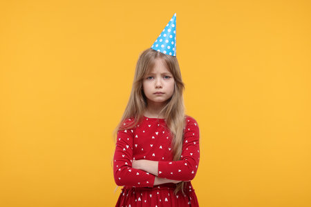 Unhappy little girl in party hat with crossed arms on yellow backgroundの写真素材