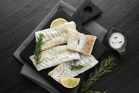 Fresh raw cod fillets, rosemary, lemon and salt on black table, flat lay