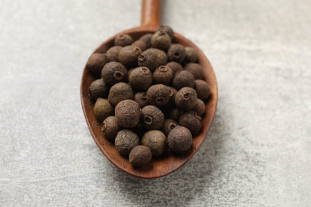 Aromatic allspice pepper grains in wooden spoon on gray table, closeup