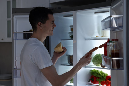 Man with cheese taking sausages out of refrigerator in kitchen at nightの写真素材