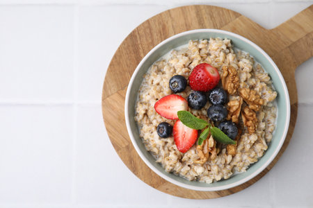 Tasty oatmeal with strawberries, blueberries and walnuts in bowl on white tiled table, top view....