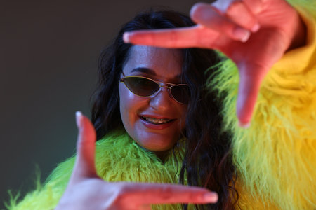 Portrait of beautiful woman in yellow fur coat and sunglasses posing on dark background with neon...