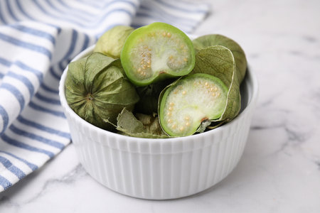 Fresh green tomatillos with husk in bowl on light marble table, closeup