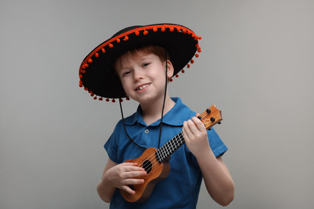 Cute boy in Mexican sombrero hat playing ukulele on grey backgroundの写真素材