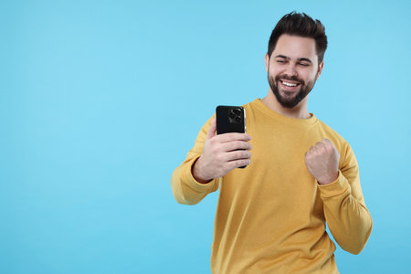 Happy young man using smartphone on light blue background, space for text