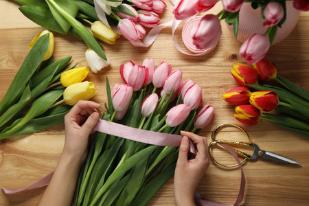 Woman making beautiful bouquet of fresh tulips and ribbon at wooden table, top viewの写真素材