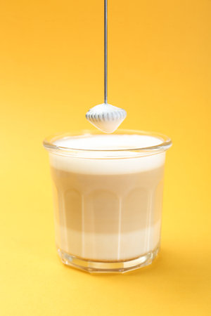 Mini mixer (milk frother) above tasty cappuccino in glass on yellow backgroundの写真素材