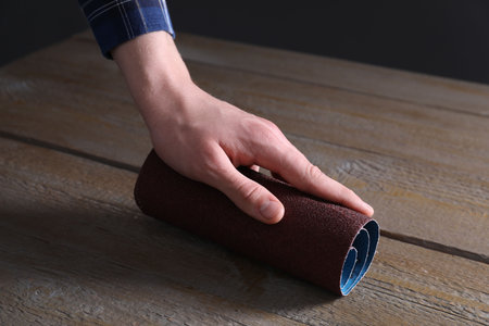 Man polishing wooden table with rolled sheet of sandpaper, closeup