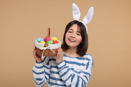 Easter celebration. Happy woman with bunny ears and wicker basket full of painted eggs on beige...