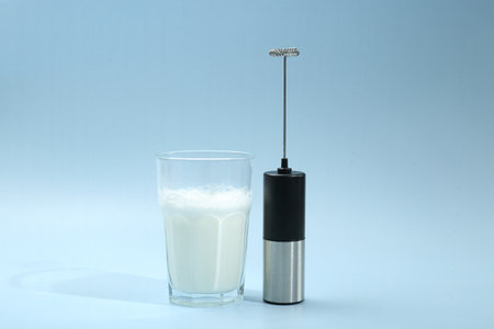 Mini mixer (milk frother) and whipped milk in glass on light blue backgroundの写真素材